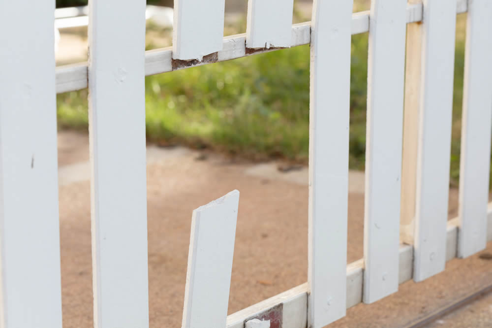 Close-up of a damaged white wooden fence with a broken vertical slat, showing wear and exposed areas against a blurred outdoor background.