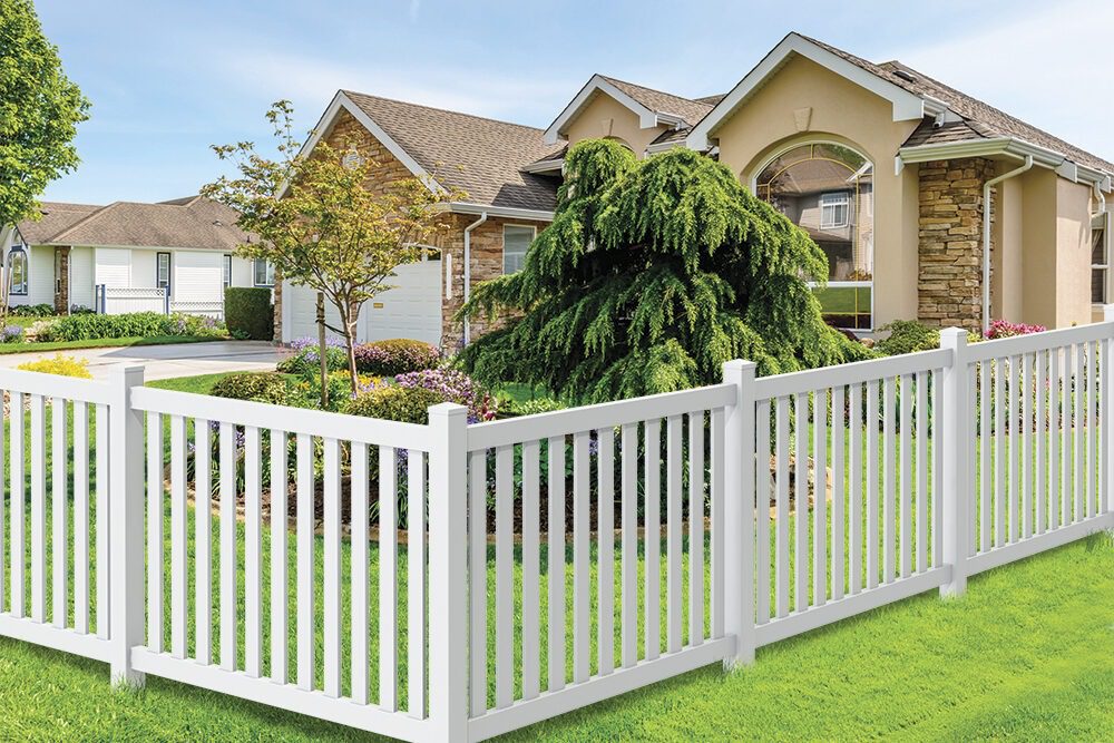 A suburban house with a stone facade, manicured lawn, landscaped garden, and a white picket fence in the foreground on a sunny day.