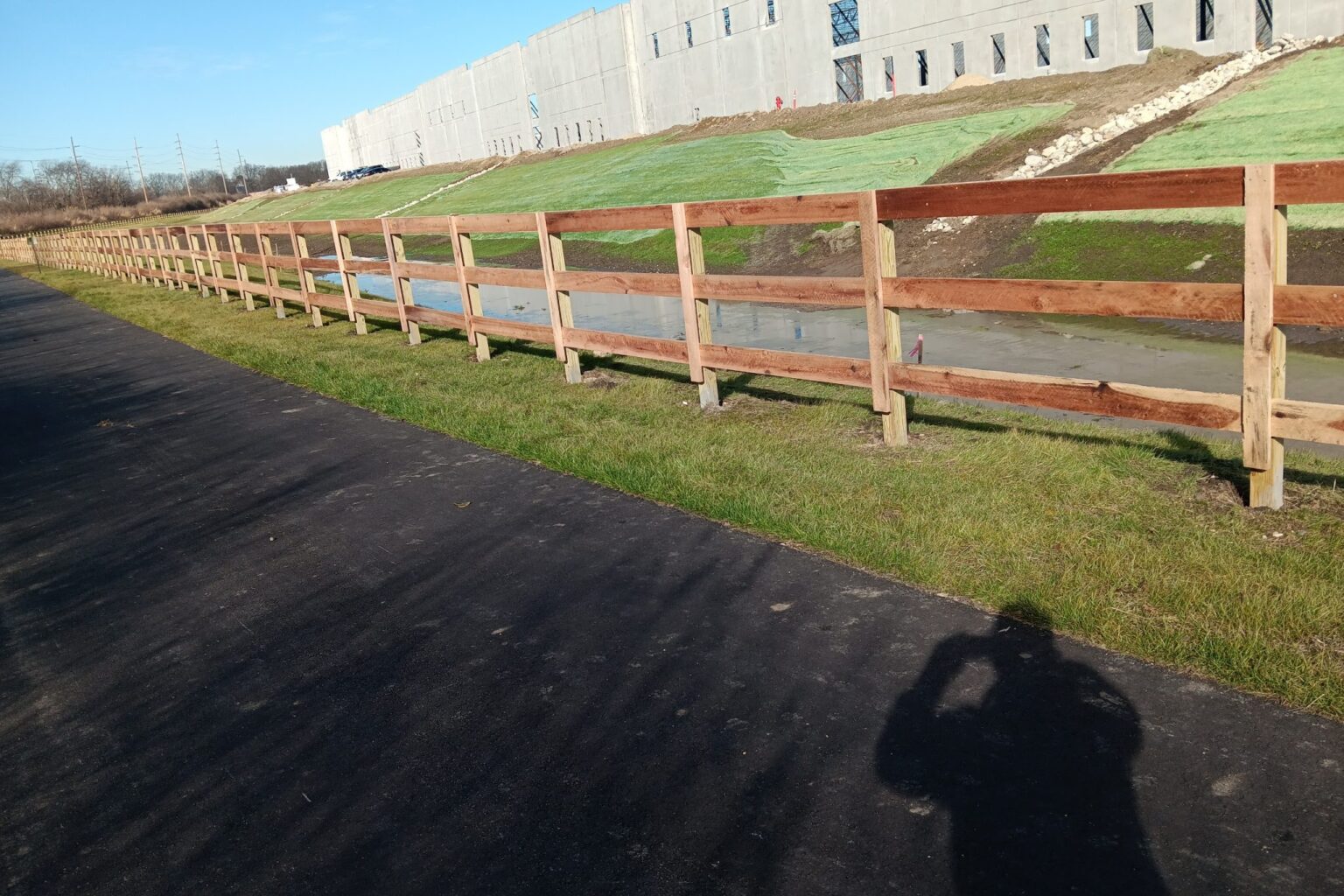 A paved trail runs parallel to a wooden fence and a grassy embankment beside a large concrete building under construction.