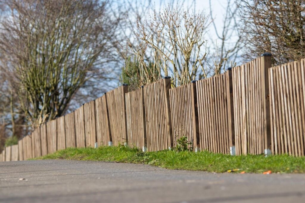 Long wooden fence along a sidewalk with trees behind it.