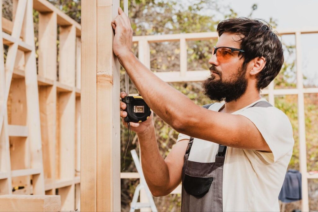 Bearded man in safety glasses measures a wooden frame with a tape measure.