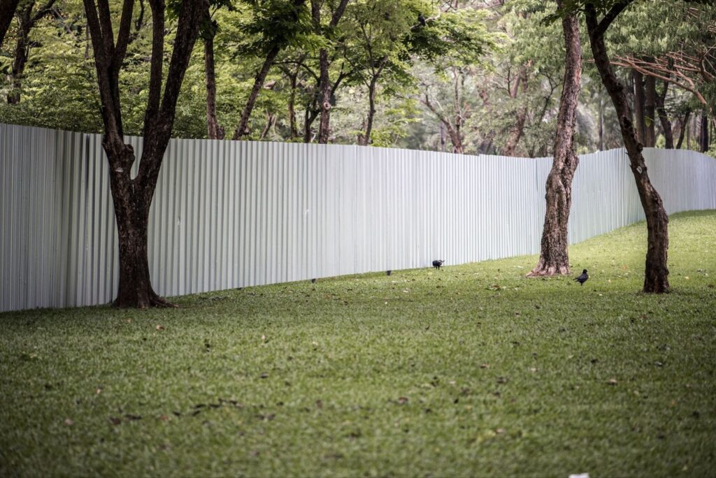 White corrugated fence curves through a grassy, tree-filled park.
