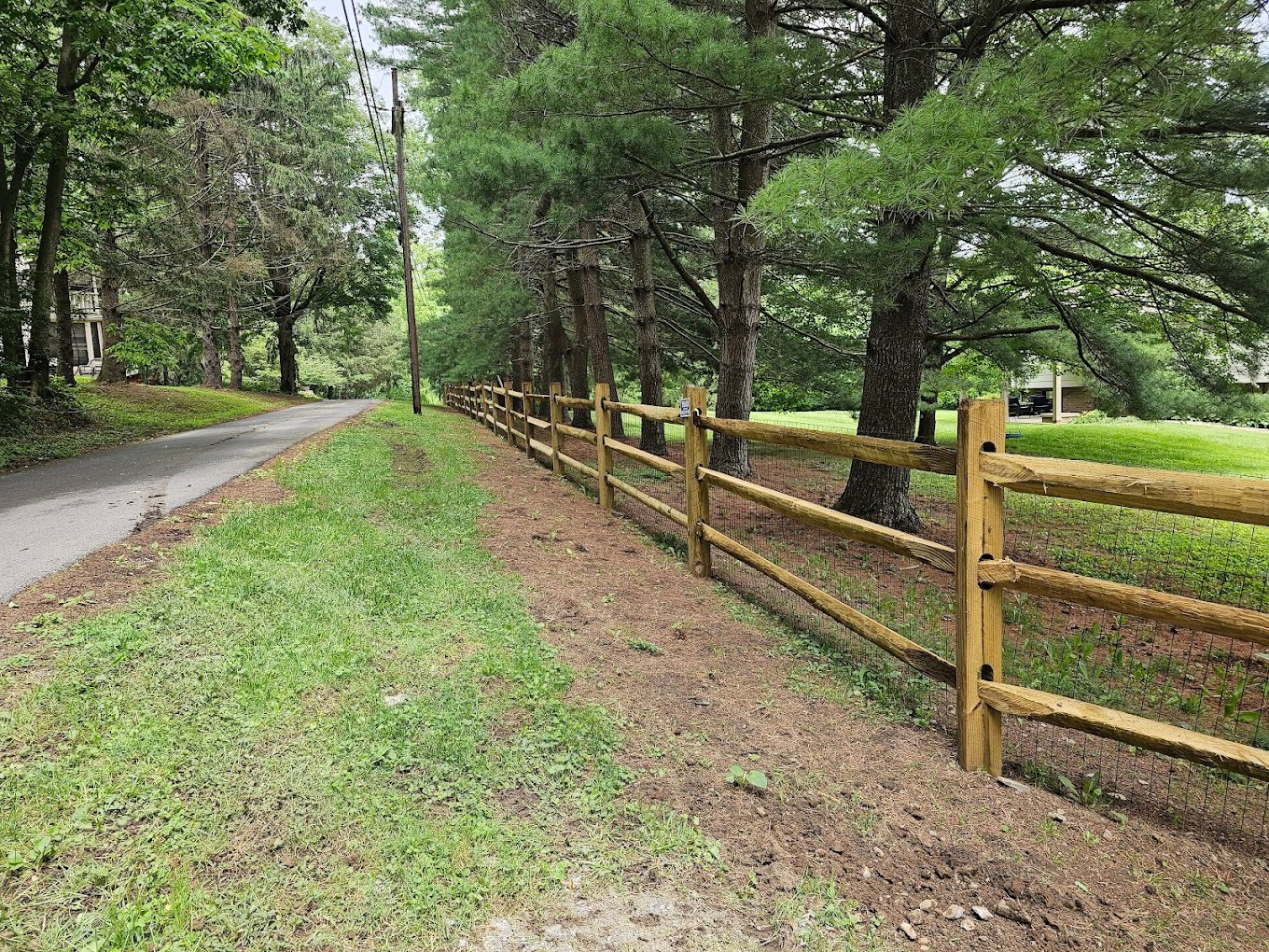 A wooden split-rail fence runs alongside a narrow paved road bordered by grass and tall trees on a cloudy day, showcasing the rustic charm often compared in vinyl vs. wood fencing choices.
