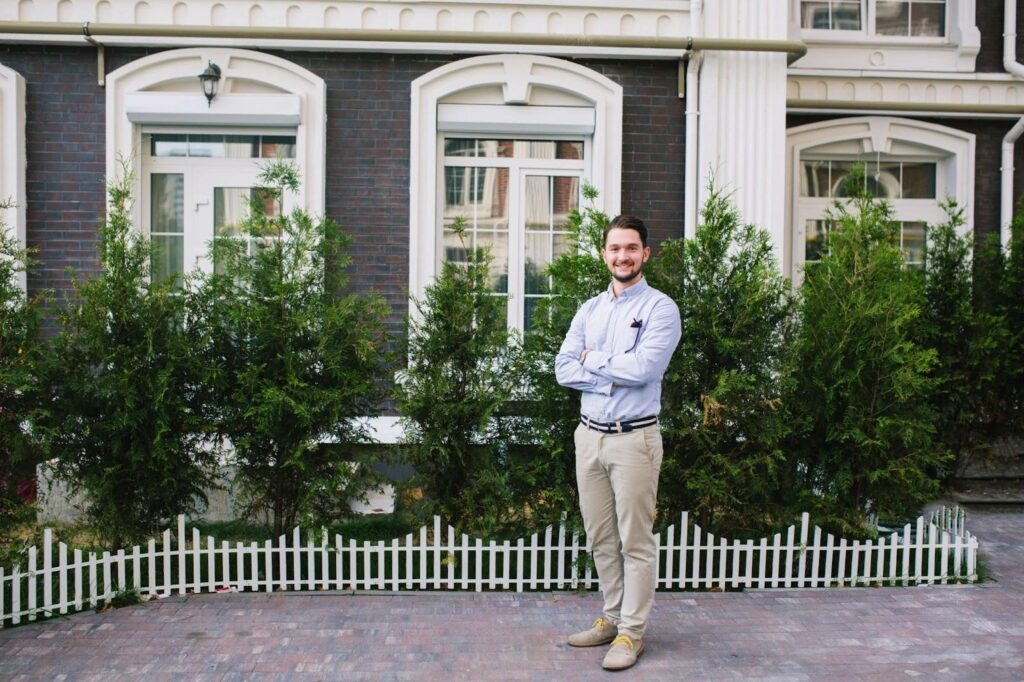 Man stands with arms crossed in front of a house and shrubs.