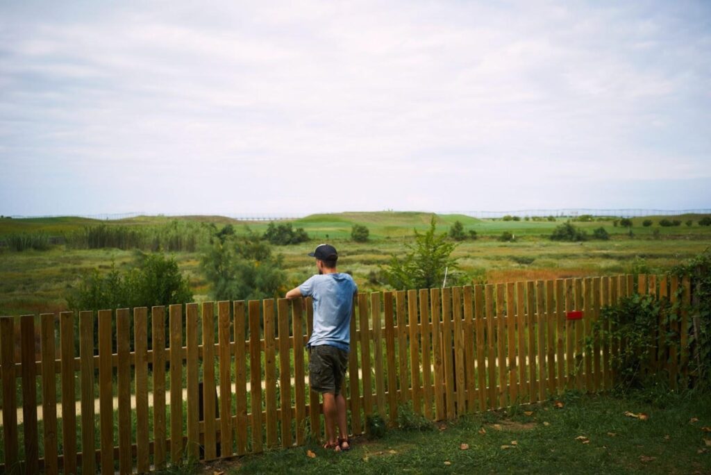 Person leans on a wooden fence, looking out over open countryside.