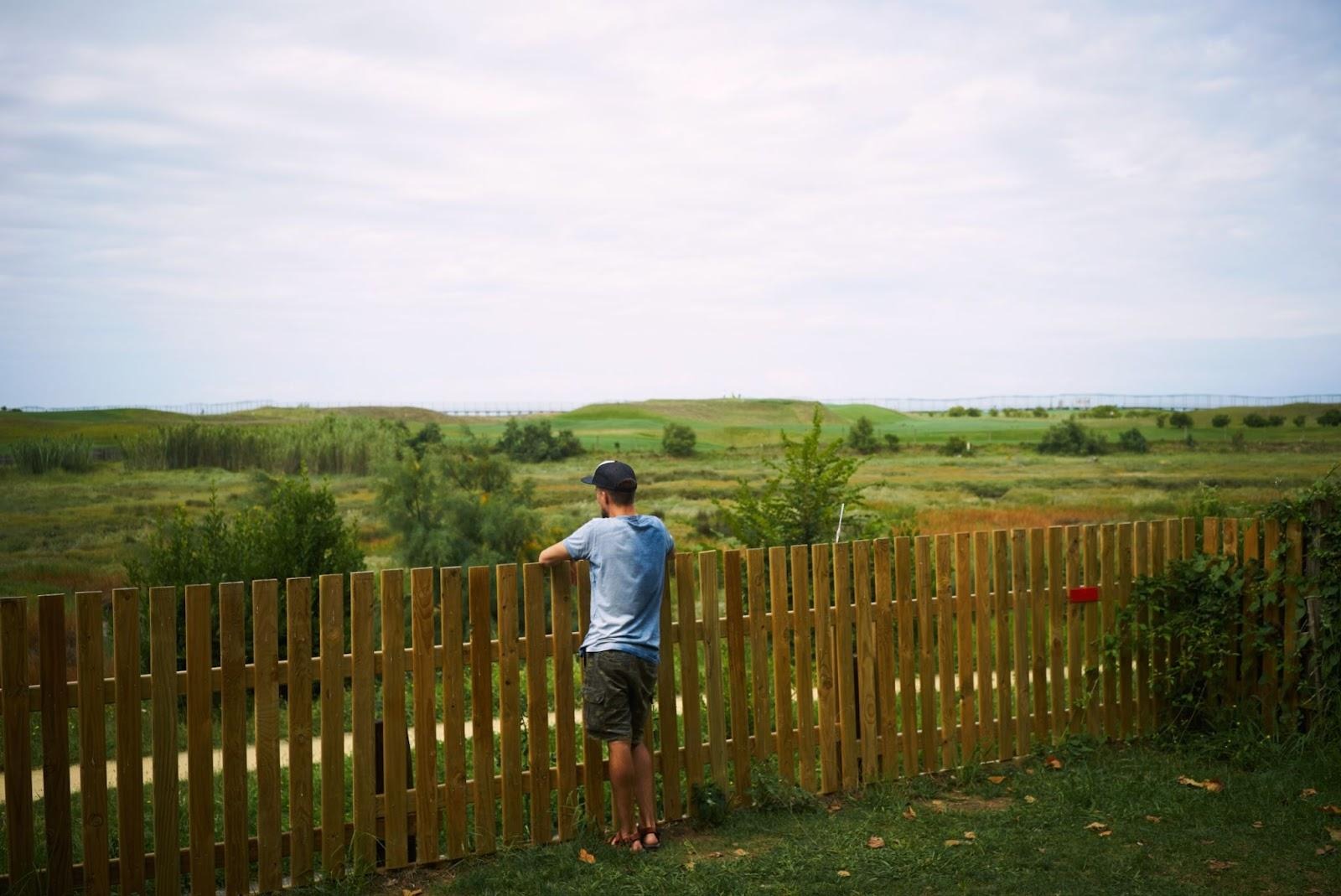 A person in a blue shirt and shorts stands by a wooden fence, gazing over the green, grassy landscape under a cloudy sky, with the fence adding rustic charm to the serene scene.