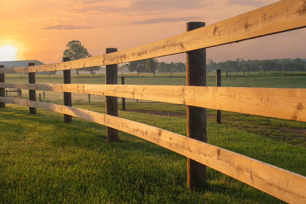 A wooden fence stretches across a grassy field at sunrise, with trees and a soft orange sky in the background.