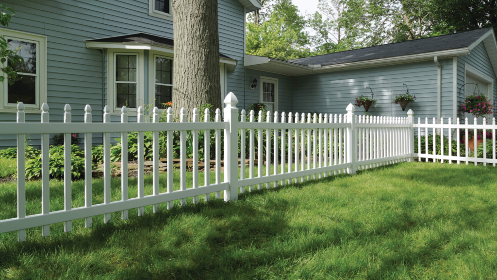 White picket fence enclosing a green backyard beside a blue house with trees and flowers.