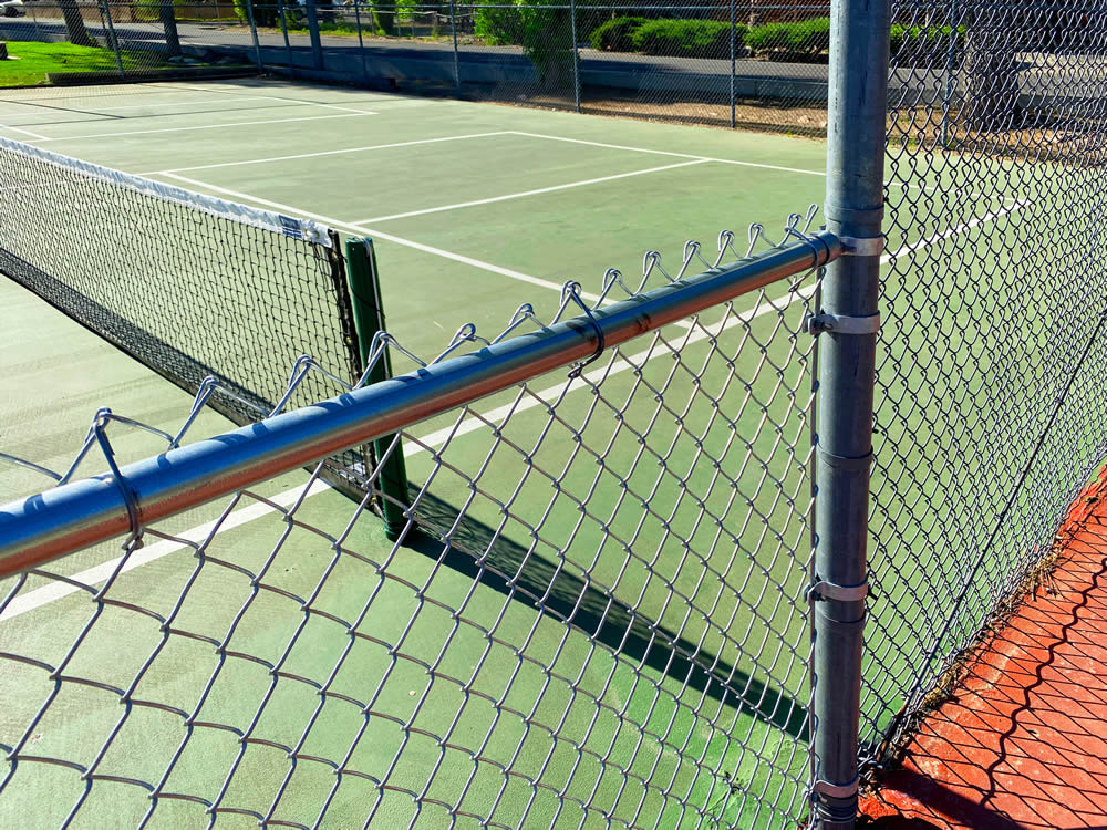 Tennis court viewed through a chain-link fence, with the net and court lines visible in bright daylight.