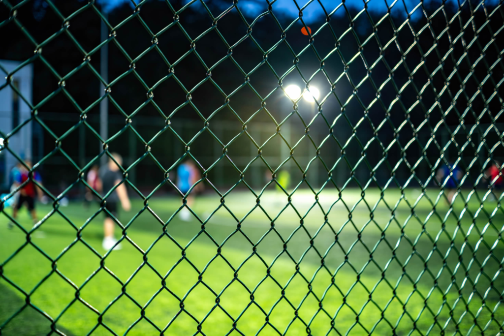Blurred night soccer game seen through a chain-link fence, with bright field lights in the background.