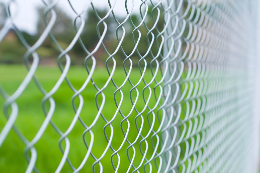 Close-up view of a sturdy metal fence, focusing on the chain-link pattern with green grass and trees softly blurred in the background.