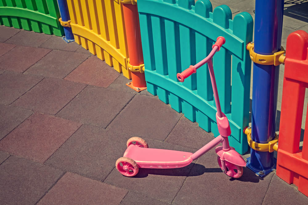 A pink three-wheeled scooter is parked on brown rubber tiles next to colorful vinyl playground fencing in an outdoor play area, creating safer play areas for children.