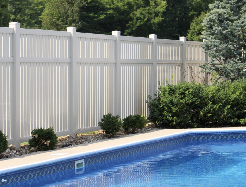 White privacy fence bordering a backyard swimming pool, with landscaped shrubs and mature trees behind the fence.